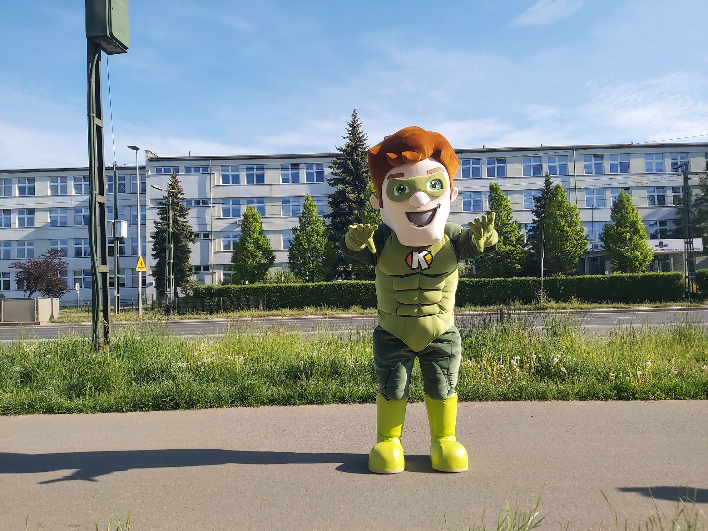Na fotografii widnieje strój reklamowy stylizowany na chłopaka w zielonych okularach pozujący do zdjęcia przed siedzibą firmy Elabika